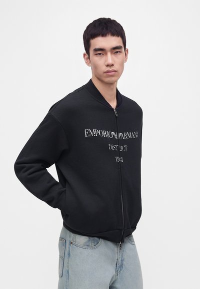 Jeune homme portant une veste zippée noire avec le texte Emporio Armani, posant avec les mains dans les poches, jean bleu clair, fond neutre.