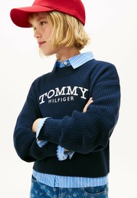 Maglione lavorato a maglia blu navy con logo bianco "TOMMY HILFIGER". Indossato sopra una camicia a strisce blu chiaro con polsini scallop. Cappello rosso indossato.