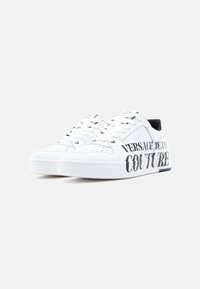 Λευκά αθλητικά παπούτσια με λεία δερμάτινη υφή. Διαθέτουν μαύρο τυπωμένο κείμενο "VERSACE JEANS COUTURE" στην πλευρά και σχέδιο με κορδόνια.