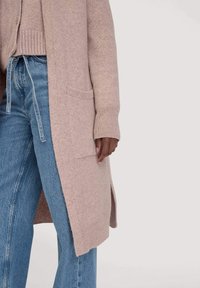 Vrouw in een beige lange cardigan met zakken, een gebreide trui en high-waisted blauwe jeans met een gestrikt riempje.
