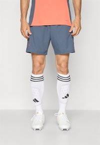 Herreur outfit til sport bestående af en koral top, grå shorts, hvide knæhøje sokker med sorte striber og hvide fodboldstøvler med designdetaljer.