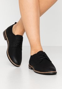 Chaussures de ville noires avec une tige en cuir lisse, des bords scallopés et un laçage. Elles présentent un petit talon avec une semelle beige clair.