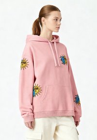Rosa hoodie med avslappnad passform, utrustad med blå och gula solbroderier på ärmarna, dragsko-huva och känguruficka.