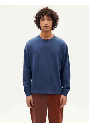 Junger Mann mit lockigem Haar, der ein langärmeliges blaues Hemd und rostfarbene Hose trägt und vor einem schlichten weißen Hintergrund steht.