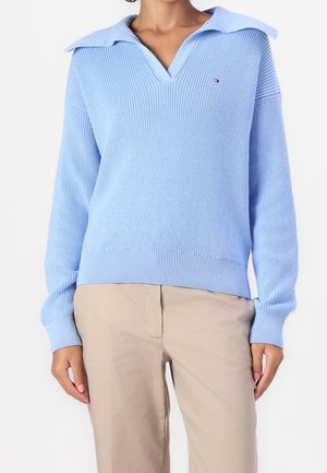 Kobieta ubrana w jasnoniebieski dzianinowy sweter z szerokim kołnierzem i beżowe spodnie, stojąca na tle jednolitego białego tła.