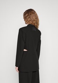 Blazer nero su misura con maniche lunghe, dettaglio a taglio nella parte posteriore e un’etichetta per la cura dei tessuti attaccata al collo. Texture morbida.