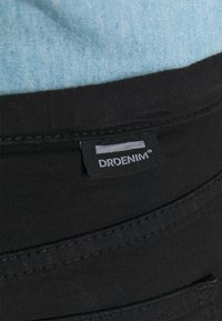 Cintura de tecido de denim preto com costura visível e uma etiqueta exibindo "DRDENIM" em branco. Denim azul claro está parcialmente visível acima.