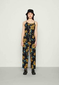 Mulher vestindo um macacão preto de perna larga com estampado floral amarelo, chapéu bucket preto, brincos de argola prateados e sapatos plataforma pretos, em pé.