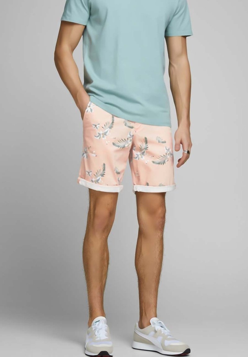 Shorts rose clair avec des motifs de feuilles de palmier vertes, présentant un ourlet roulé. Associés à un t-shirt bleu clair et des baskets blanches.