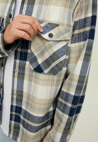 Chemise à boutons en plaid beige, marine et blanche. Caractéristiques d'une poche poitrine, de motifs contrastés et d'une coupe décontractée avec des poignets boutonnés.