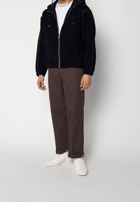 Schwarze Zip-Jacke aus strukturiertem Stoff, mit Kapuze und elastischen Manschetten, kombiniert mit dunkelbraunen Hosen und weißen Sneakern. Einfaches Design.