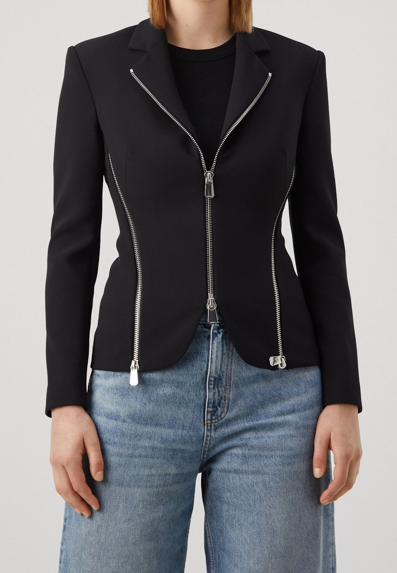 Femme portant un blazer noir cintré avec plusieurs fermetures éclair argentées, associé à un jean denim bleu clair, debout devant un fond uni.