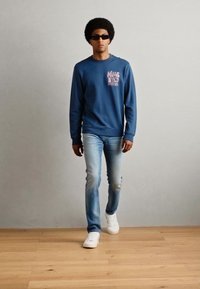 Felpa blu con una grafica stampata, abbinata a jeans azzurri e sneakers bianche. Il modello indossa occhiali da sole scuri e ha un'acconciatura afro.