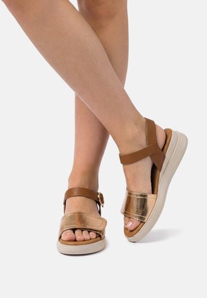 Beine mit offenen Sandalen, die metallisch roségoldfarbene Vorderriemen, braune Knöchel- und Fersenriemen sowie weiße, gepolsterte Sohlen auf weißem Hintergrund tragen.