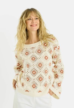 Mujer con cabello rubio ondulado que lleva un suéter de crochet crema con patrones de diamantes naranjas y pantalones blancos, sonriendo con las manos en los bolsillos.