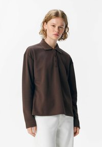 Braunes Langarm-Poloshirt mit Kragen, ausgestattet mit einer Knopfverschluss, entspannter Passform und glatter Textur. Kombiniert mit weißen Hosen.