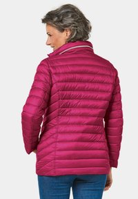 Magenta pufferjack met een hoge kraag, geborduurd textuur en getailleerd ontwerp. Heeft een ritsaccent en is gecombineerd met blauwe jeans.