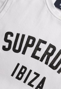Top sin mangas de algodón blanco que presenta un audaz texto en brillante negro que dice "SUPERDRY IBIZA". Textura suave con un clásico escote redondo.
