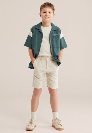 Chlapec stojaci s rukami v priehlbniach v džínsovej tyrkysovej košeli s krátkym rukávom cez biele tričko, béžové šortky, biele ponožky a béžové tenisky.