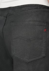 Zoom na zadnji žep črnih jeans hlač z diskretno vezeno valovito obliko in malim rdečim vezenim detajlom na robu žepa.