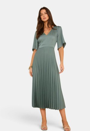 Vrouw draagt een lichtgroene geplooide midijurk met korte mouwen en V-hals, gecombineerd met gouden armbanden en beige sandalen met bandjes.