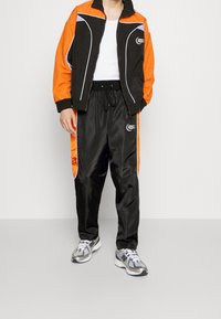 Black and orange athletic jacket with white accents, paired with black satin pants featuring orange side stripes and a logo. Gray sneakers complete the look.
