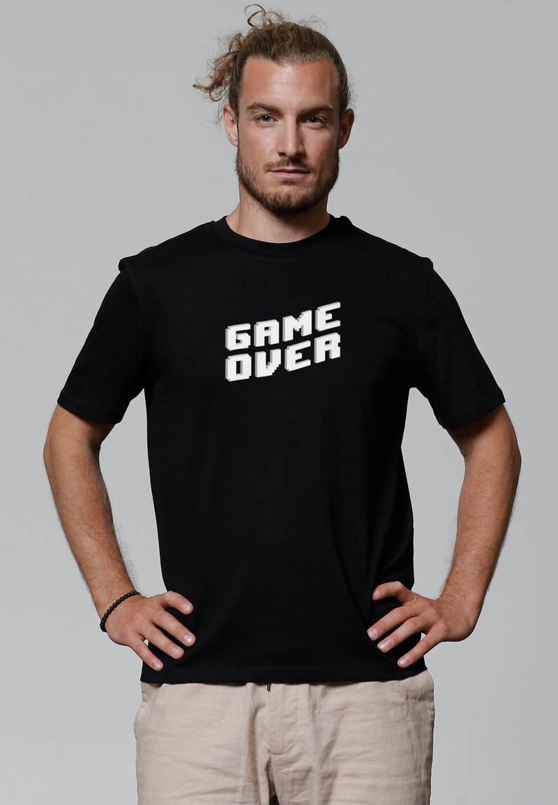 Schwarzes T-Shirt mit weißem pixeliertem Text, der "GAME OVER" sagt. Kurze Ärmel und Rundhalsausschnitt, aus Baumwollstoff gefertigt.