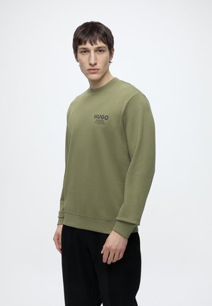 Jeune homme portant un sweat Hugo vert olive et un pantalon noir, debout devant un fond clair uni.