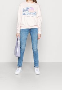Ljusrosa sweatshirt med våg- grafik och texten "DEN STORA VÅGEN," i kombination med ljusblå jeans och vita sneakers, medan hon håller en lavendelfärgad handväska.