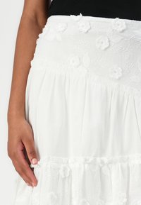 Vit broderad kjol med blommönster, volangkant, texturerat tyg och resårmidja. Hand som håller i kjolens sida.