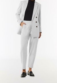 Blazer gris claro con botones de doble botonadura, corte entallado. Combinado con pantalones gris claro a juego y mocasines de charol negros.