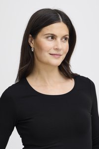 Donna con lunghi capelli scuri che indossa orecchini a cerchio d'argento e una maglietta nera con scollo rotondo, guarda leggermente verso destra su uno sfondo uniforme.