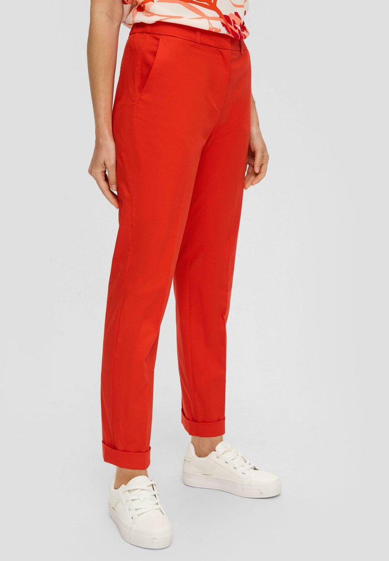 Rode broek met een getailleerde pasvorm, voorzien van zijzakken en omgeslagen manchetten. Gepaarde met witte sneakers, wat een casual uitstraling benadrukt.