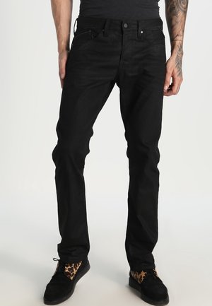 Homme portant un jean noir slim et des chaussures noires avec des accents à imprimé léopard, debout les mains détendues le long du corps.