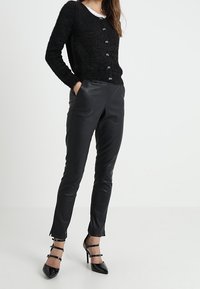 Musta pörröinen neuletakki hopeisilla napeilla, yhdistetty istuviin mustiin nahkahousuihin. Kiiltävät mustat korkeakorkoiset kengät nilkkaremmeillä.