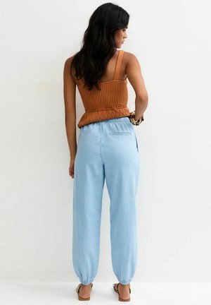 Woman with long dark hair wearing a sleeveless brown knit top and light blue elastic ankle pants, standing against a white wall.