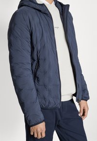 Man draagt een marineblauwe gewatteerde jas over een crèmekleurige trui en marineblauwe broek, staand tegen een witte achtergrond met ontspannen handen.