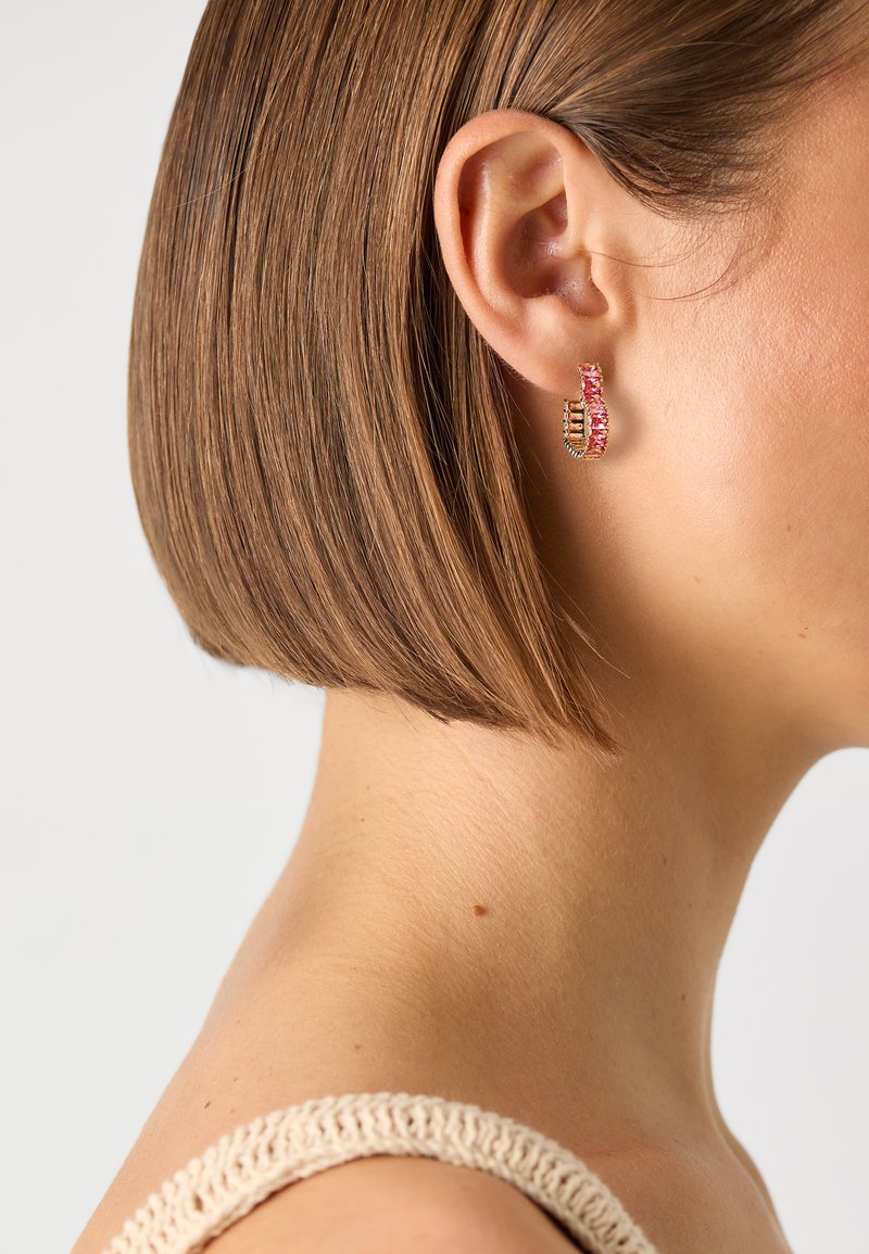 Auksinės spalvos auskarai-žiedai su rožiniais ir skaidriais akmenimis, išryškinantys bangos dizainą su pakaitomis besikeičiančiomis spalvomis ir poliruotu paviršiumi.