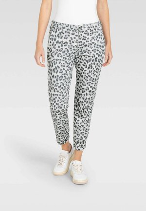 Femme portant un pantalon blanc et gris imprimé léopard et des baskets blanches, debout sur un fond clair.