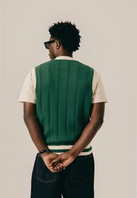 Hombre con cabello rizado oscuro que lleva gafas de sol negras, un chaleco tejido verde sobre una camisa beige y jeans oscuros, de pie con las manos entrelazadas detrás de la espalda.