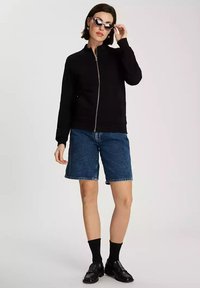 Czarna kurtka zapinana na zamek z długimi rękawami, w połączeniu z niebieskimi szortami dżinsowymi. Dopełnieniem są okulary przeciwsłoneczne w kocim kształcie oraz czarne skarpetki z butami.