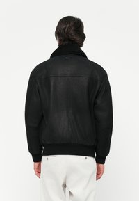 Black bomber jacket with textured fabric, wide collar, and ribbed cuffs. Features a logo on the back and a fitted waistband.