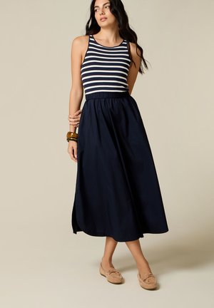 Vrouw met een marineblauw-wit gestreepte mouwloze top, een marineblauwe maxirok, beige loafers en armbanden, staand tegen een effen achtergrond.