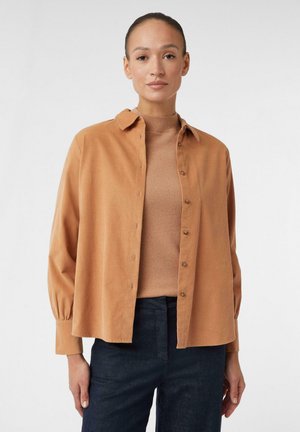 Camicia con bottoni in arancione bruciato, realizzata in tessuto morbido. Presenta un colletto classico, polsini aderenti e una silhouette rilassata. Abbinata a un maglione beige.