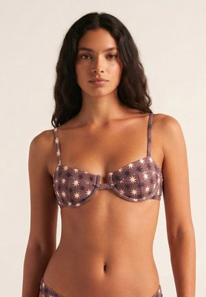 Mujer con cabello largo y oscuro que lleva un top de bikini malva con patrones de estrellas blancas sobre un fondo neutro.