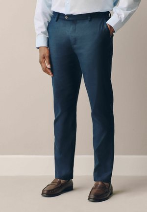 Mann trägt marineblaue Anzughose, braune Loafers und ein hellblaues Hemd mit Knöpfen, steht mit einer Hand in der Hosentasche.