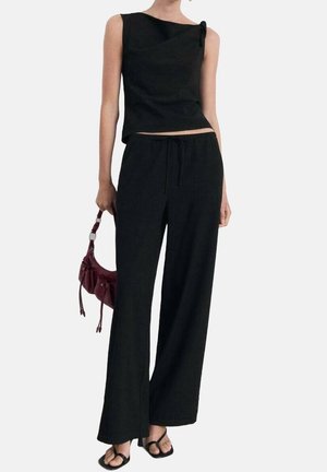 Frau trägt schwarzen ärmellosen Oberteil und weite Hosen, hält eine kleine burgunderrote Handtasche und trägt schwarze Sandalen mit offenem Zehenbereich.