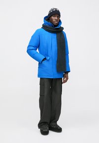 Blue puffer jacket with a high collar and zip closure, paired with a dark gray scarf. Loose-fitting dark cargo pants and black shoes.