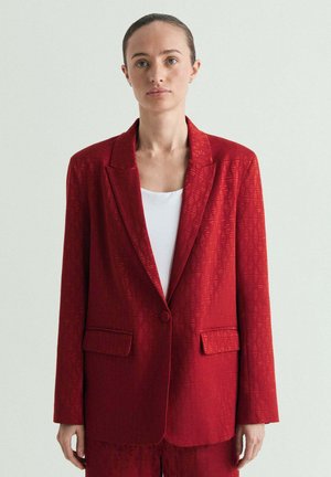 Mujer de piel pálida y ojos verdes que lleva un blazer rojo con textura sobre una camiseta blanca, de pie frente a un fondo claro y liso.