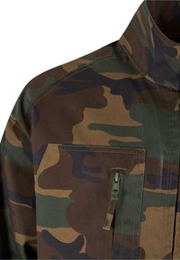 Terepmintás ing tartós anyagból; zöld, barna és fekete árnyalatokkal; elöl cipzárral záródó zsebbel és merevített gallérral rendelkezik.